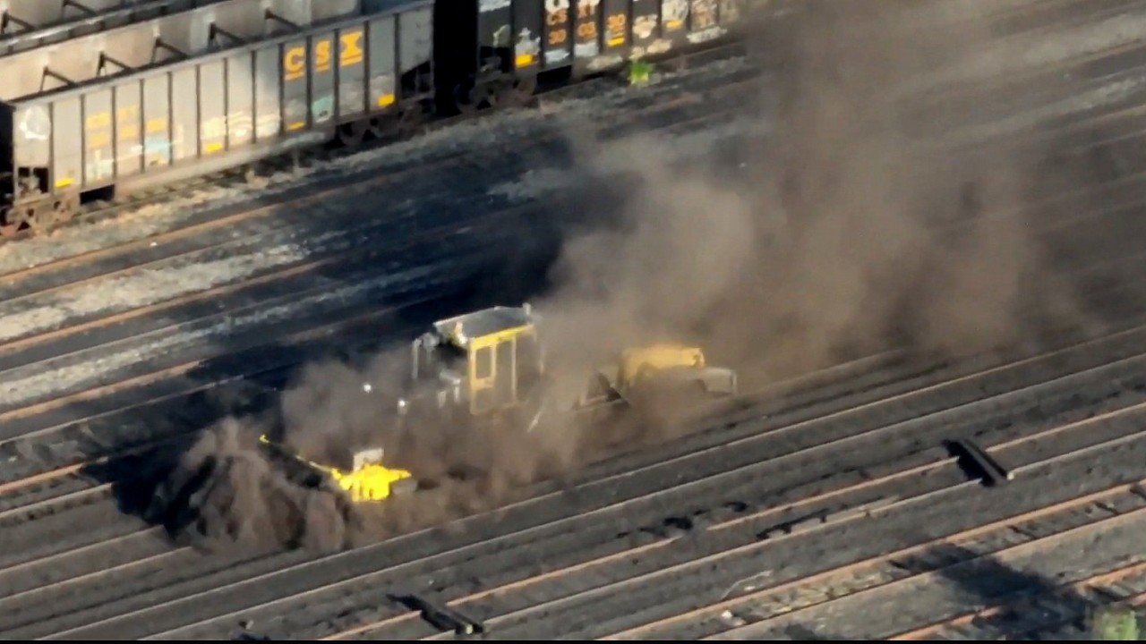 A large piece of yellow equipment on several rail lines enveloped by a huge cloud of dark dust