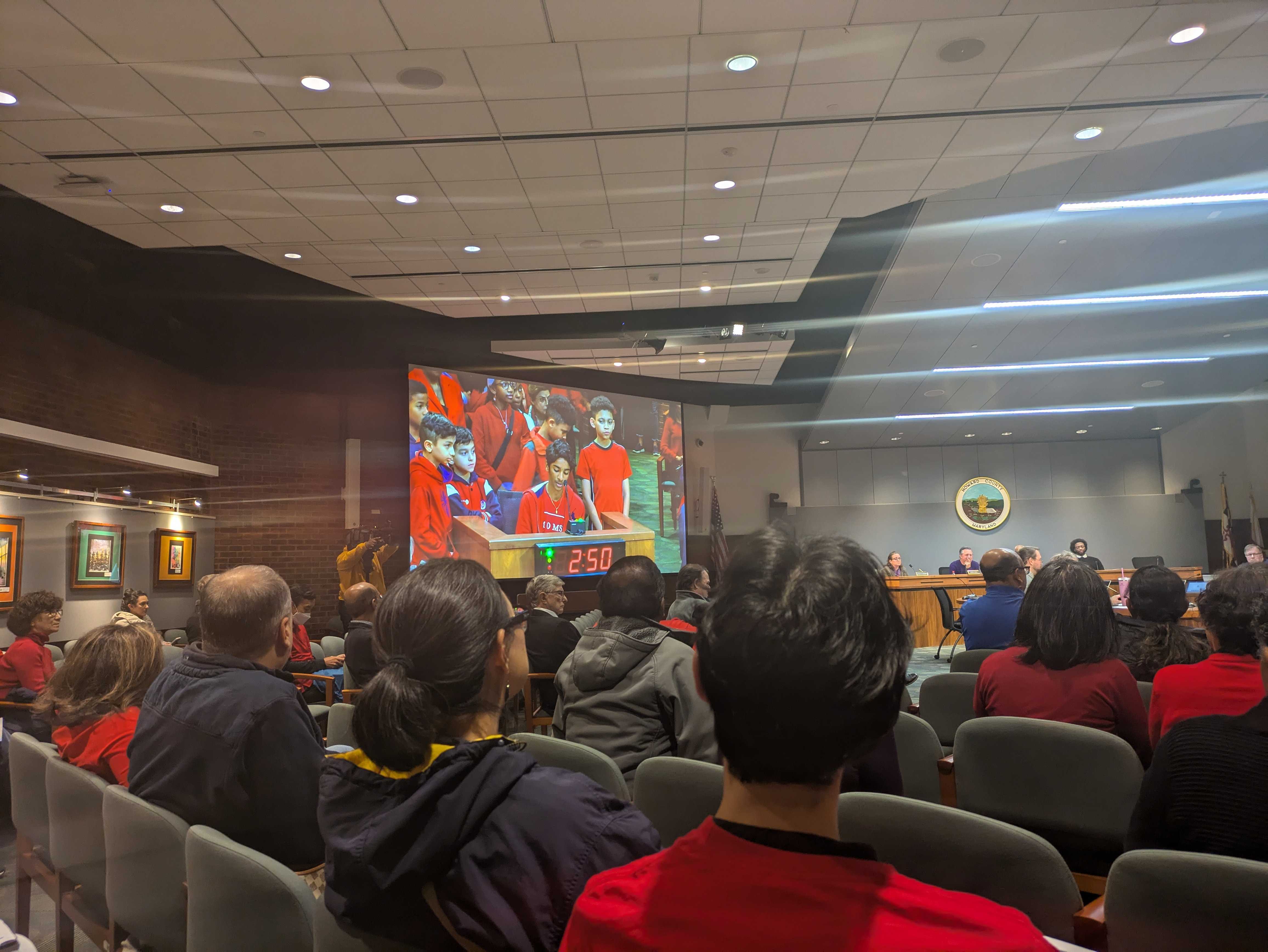 A large government meeting room from behind full of people wearing red shirts; a group of students wearing red are visible on a screen at the front of the room testifying at a podium.