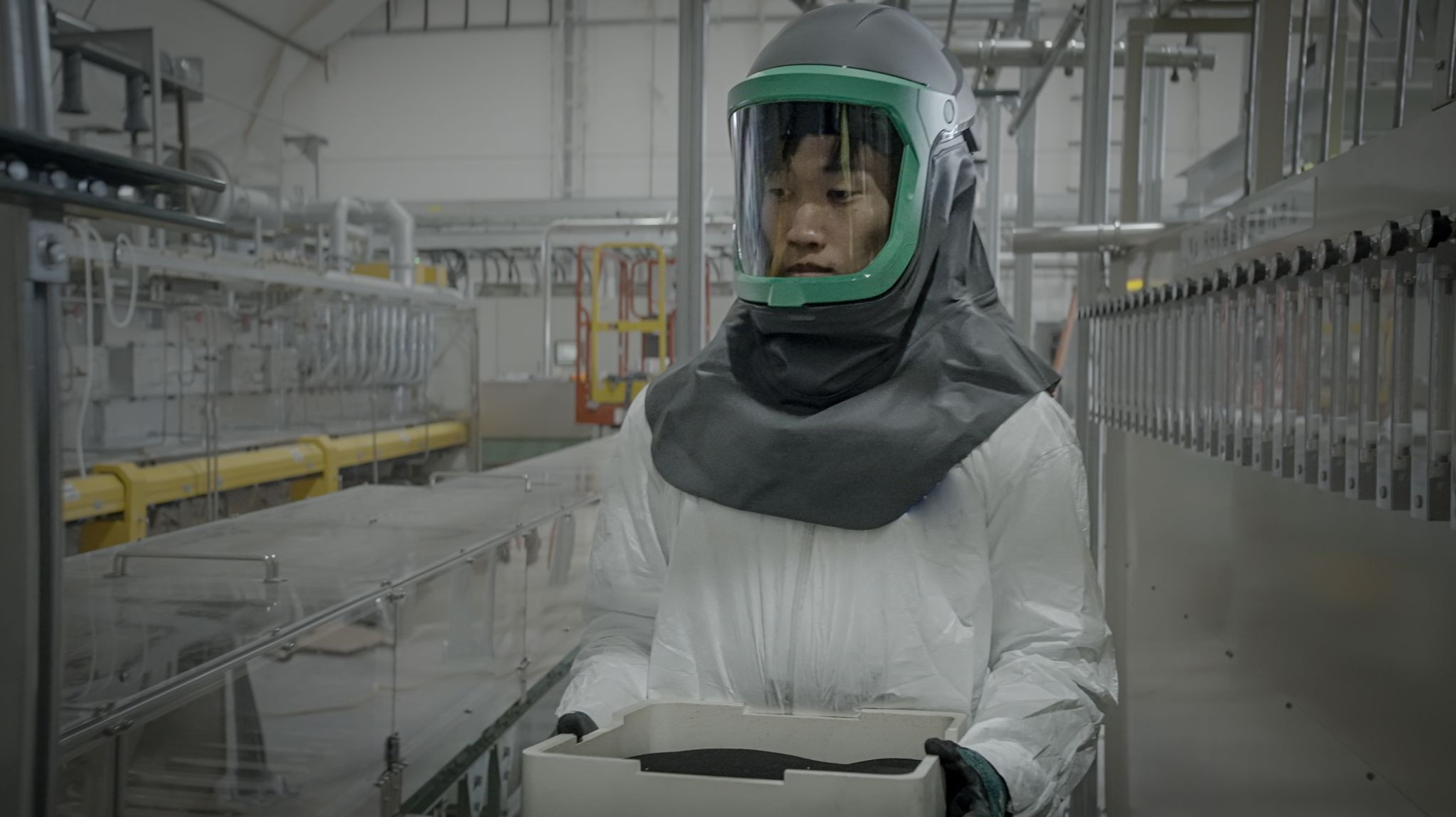 Worker in a full-coverage safety suit carrying a tray of CAM