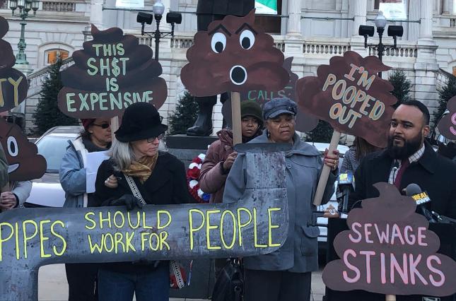 A crowd of people rallying outside of Baltimore City Hall, with signs like "Pipes should work for people" and "sewage stinks"