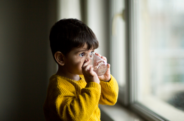 Child looking out of a window, drinking water from a glass.