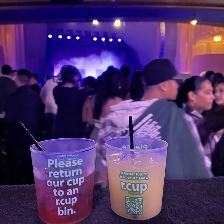 Reusable cups in front of concertgoers with printed text "Please return our cup to an r.cup bin"