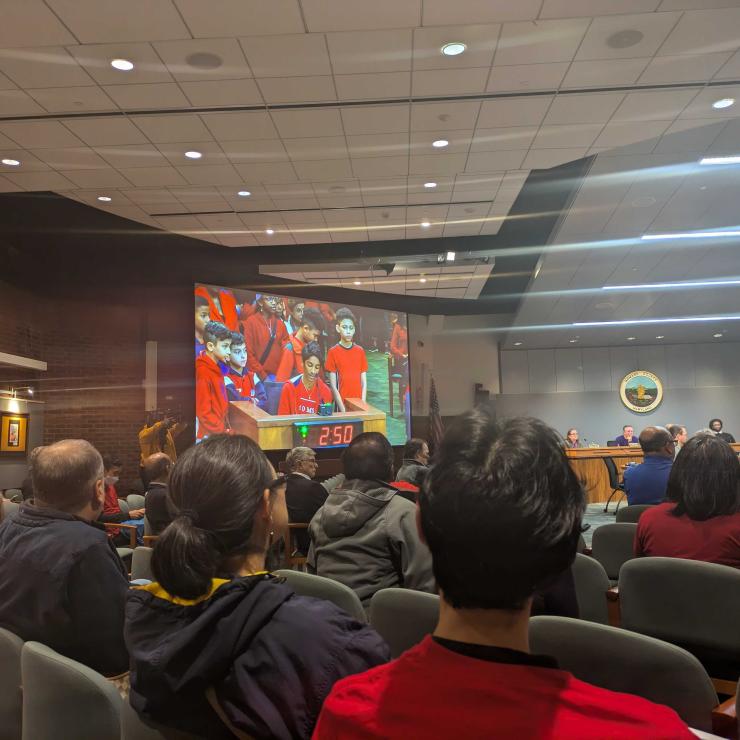 A large government meeting room from behind full of people wearing red shirts; a group of students wearing red are visible on a screen at the front of the room testifying at a podium.