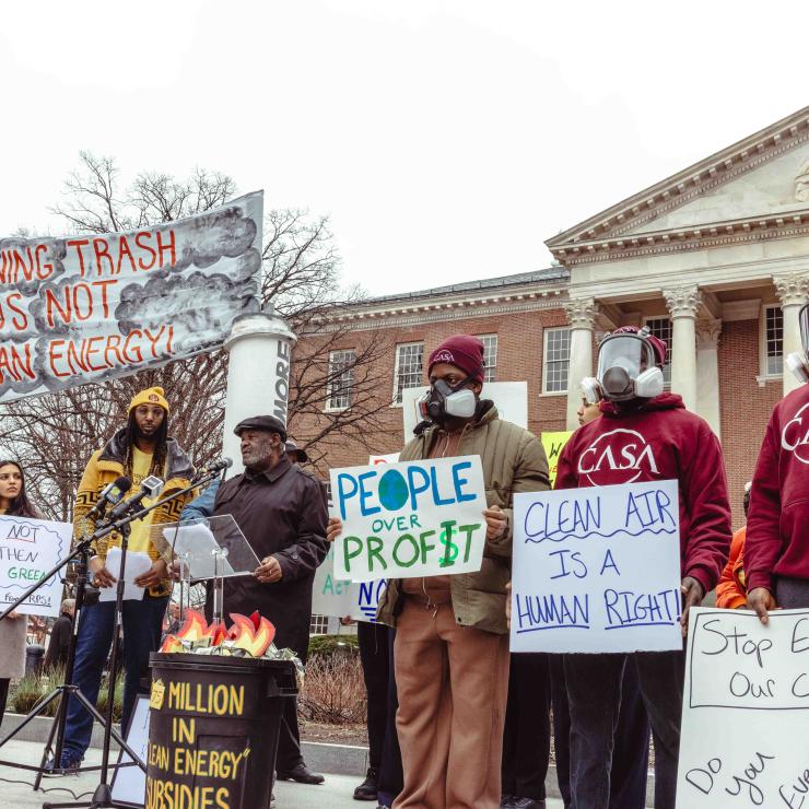 People rallying in front of the MD State House, some wearing gas masks, with signs like "Burning trash is not clean energy," "clean air is a human right," and "people over profit."