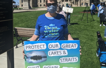 Woman outside Michigan Capitol wearing clean water action shirt and sign: "Protect our Great Lakes & Straits - Shut Down Line 5 Now"