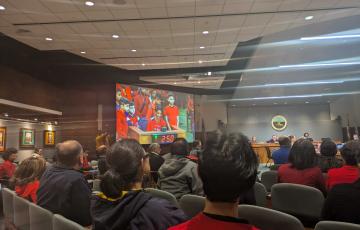 A large government meeting room from behind full of people wearing red shirts; a group of students wearing red are visible on a screen at the front of the room testifying at a podium.