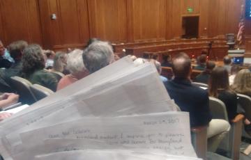 The public hearing room, full, with handwritten letters held in the foreground