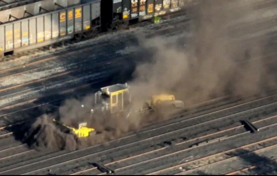 A large piece of yellow equipment on several rail lines enveloped by a huge cloud of dark dust