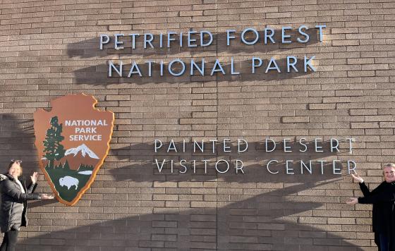 Two women posing by the sign for the Painted Desert Visitor Center at Petrified Forest National Park