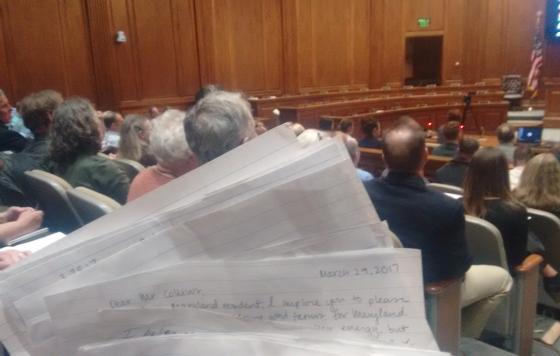 The public hearing room, full, with handwritten letters held in the foreground